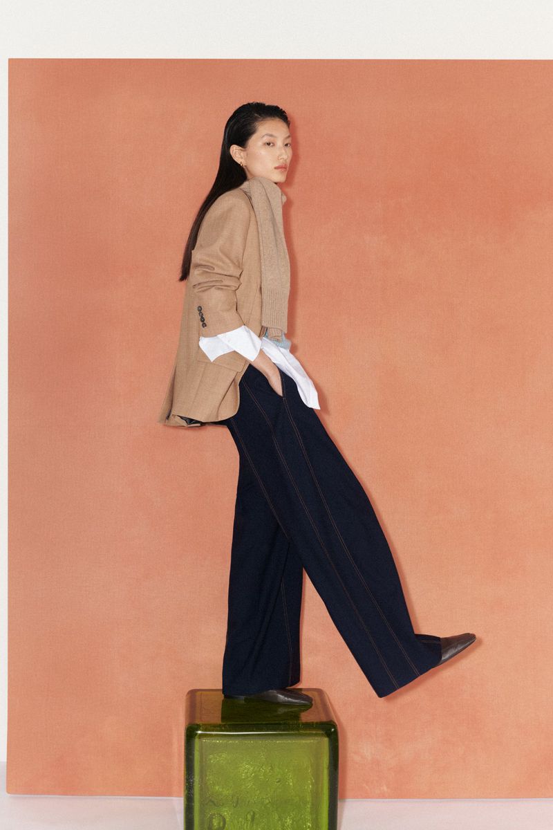Fashionable Asian woman in a tan blazer and blue trousers, strikes a pose standing atop a green block against an orange backdrop.