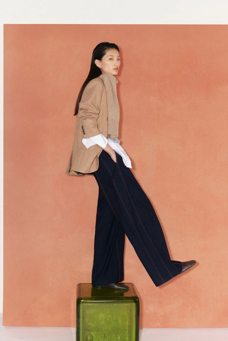Model Xinyue Guo posing on a green glass cube wearing a camel blazer and wide-leg trousers against a terracotta backdrop.