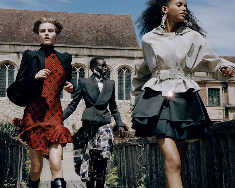 Three models wearing tailored jackets and plaid skirts walk across an outdoor bridge in front of a historic stone building.