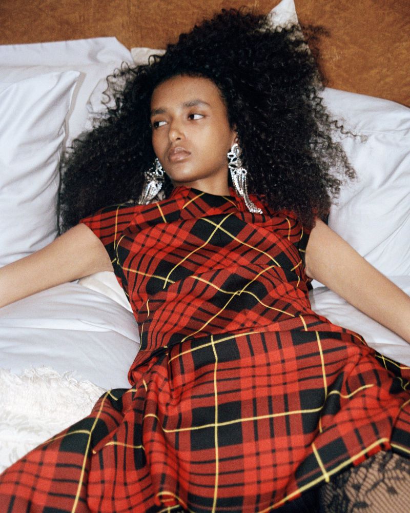 Woman with voluminous afro poses in a red tartan dress on a bed with white pillows. She wears large silver earrings.