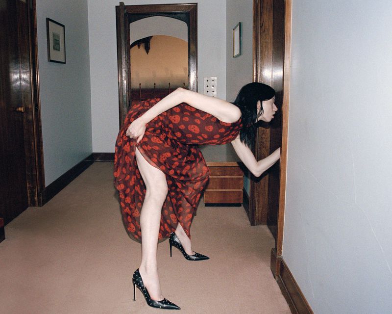 Woman in a red dress and high heels peeks around a door in an interior hallway setting with light walls and dark wood trim.