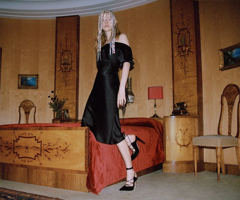 Fashionable woman in an off-the-shoulder black dress and silver headpiece, posing indoors with dark decor and vintage furniture.
