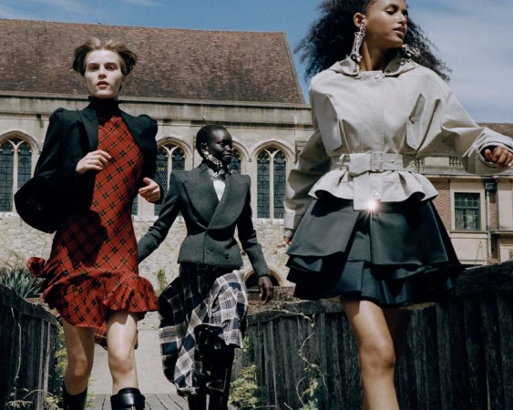 Three models wearing tailored jackets and plaid skirts walk across an outdoor bridge in front of a historic stone building.