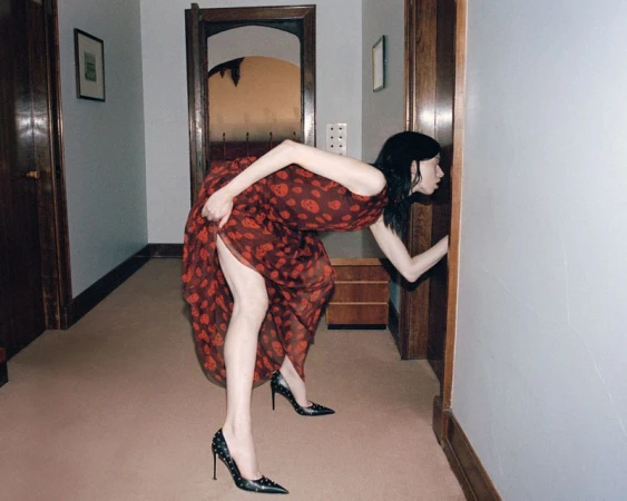 Model in a patterned red dress and black studded heels bending forward to look into a doorway in a hallway.