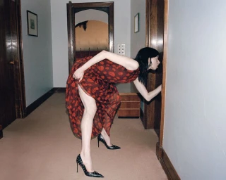 Model in a patterned red dress and black studded heels bending forward to look into a doorway in a hallway.