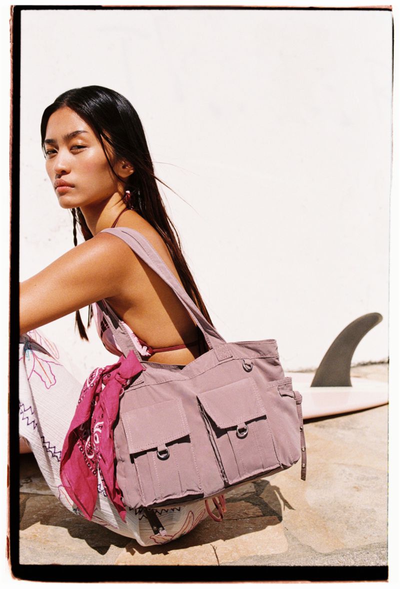 Woman poses with a mauve-colored tote bag outdoors, sitting near a surfboard, under bright sunlight, in a fashion editorial style.