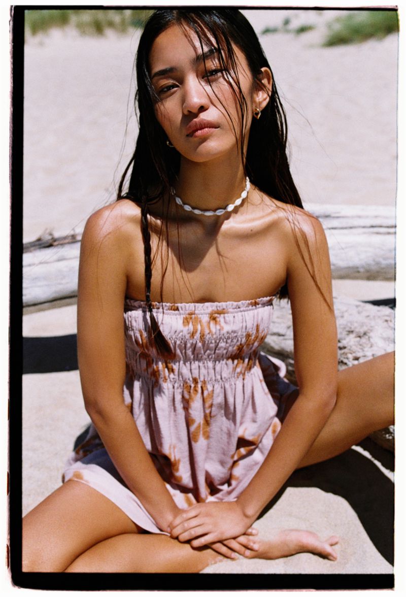 Young woman with long dark hair poses in a strapless floral dress on a sandy beach. Casual fashion, natural light, and a serene expression.