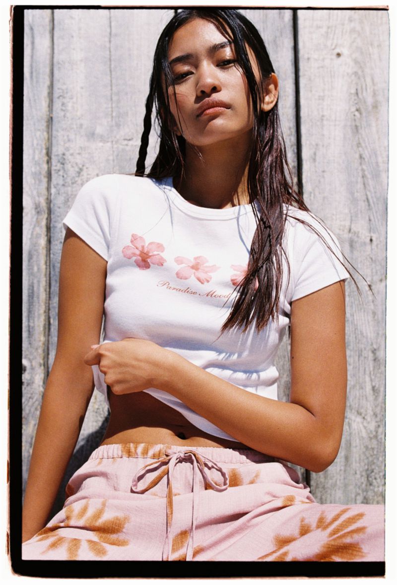 Young woman with wet hair poses in white floral t-shirt and pink tie-dye pants against a wooden background, showcasing casual fashion.