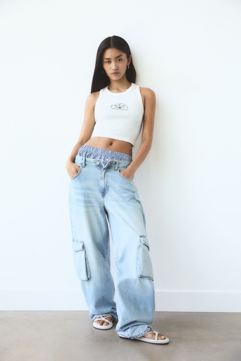 Full shot of a young Asian woman in casual fashion, featuring a white tank top and baggy light blue jeans, standing against a white wall.