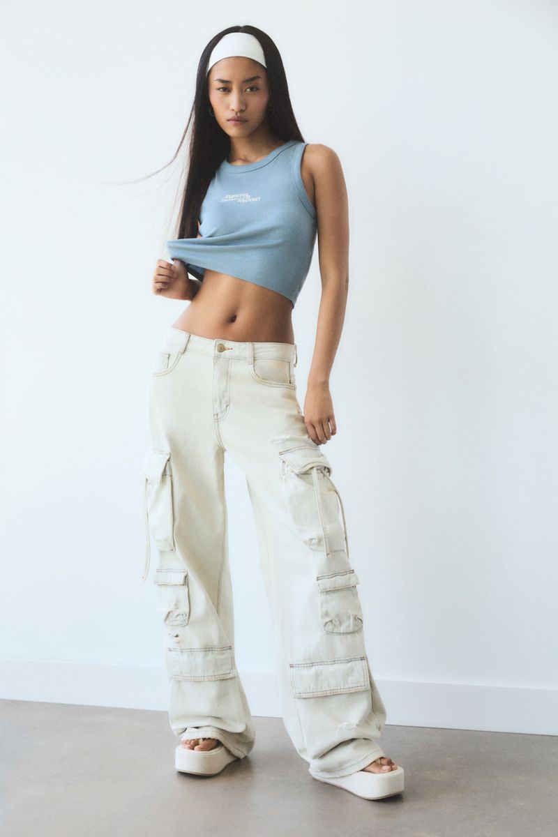 Full-length shot of a young woman wearing a blue tank top, wide-leg cargo pants, and white platform sandals posing in a brightly lit photo studio.