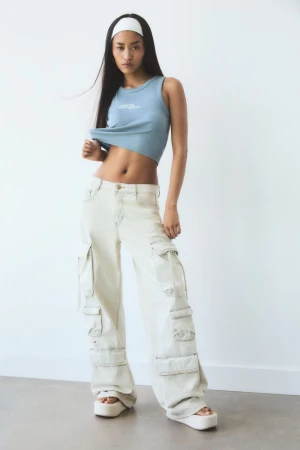 Full-length shot of a young woman wearing a blue tank top, wide-leg cargo pants, and white platform sandals posing in a brightly lit photo studio.