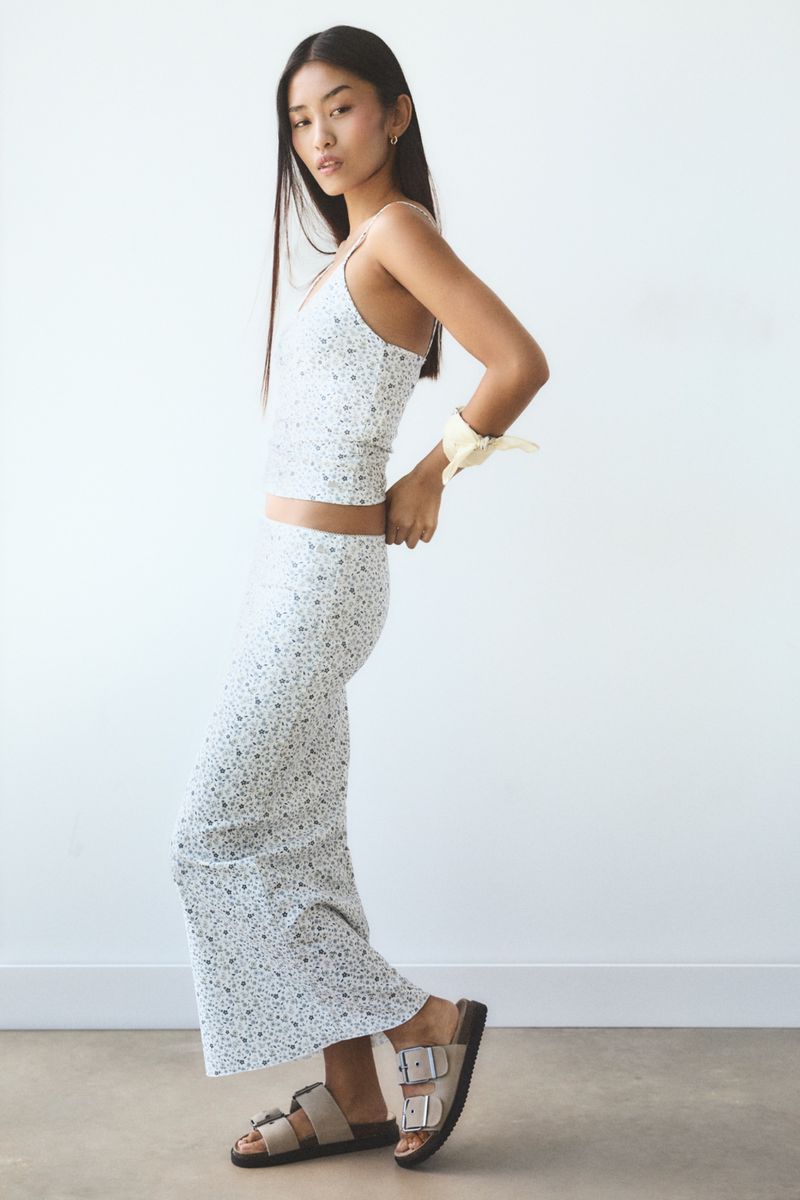 Young woman in floral print top and maxi skirt stands in front of a white wall in a studio setting, wearing sandals.