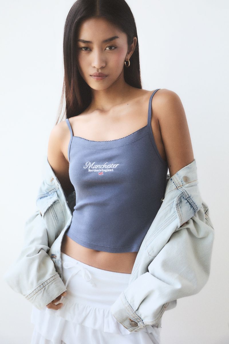 A young Asian woman poses indoors wearing a grey tank top with "Manchester Northern England" printed on it and a light denim jacket.