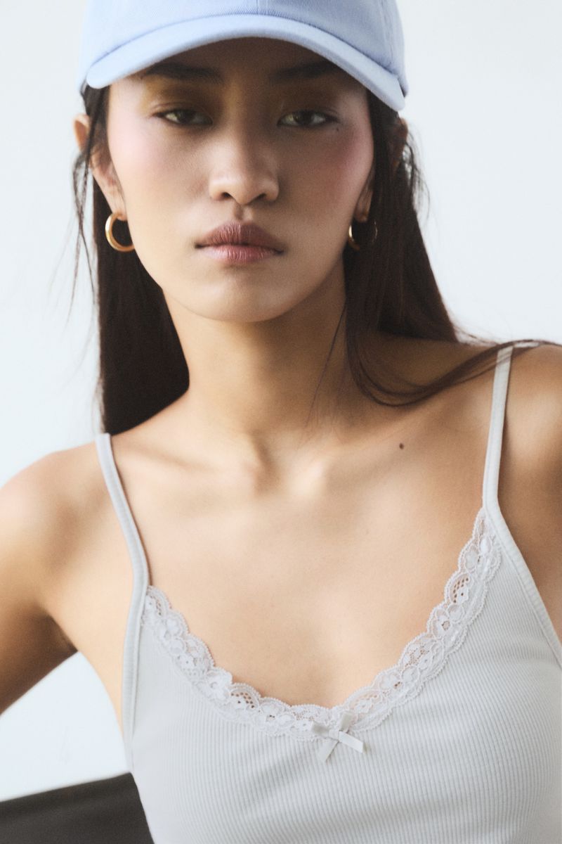 Close-up of a woman wearing a light blue baseball cap and a gray camisole with lace trim, posing against a neutral background.