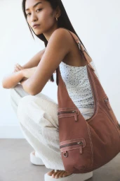 Model squatting while wearing a patterned top and white trousers with a large brown shoulder bag in a studio setting
