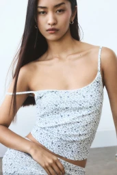 Model with long dark hair wearing a white floral camisole top against a plain studio background.