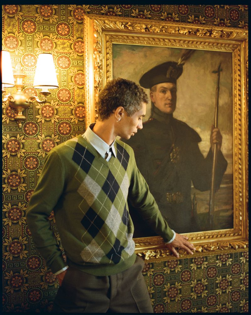 Man in argyle sweater stands in ornate room, gazing at a portrait in a gold frame, illuminated by a wall-mounted lamp. 