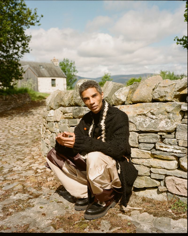 Adult male with salt-and-pepper hair squatting in countryside wearing a black knit cardigan, cream-colored pants and chunky black loafers.