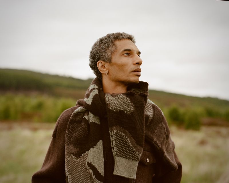Portrait of a Black adult male with graying hair, wearing a brown patterned coat and scarf outdoors in a natural setting.