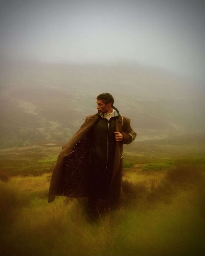 An adult male wearing a dark coat stands in a field of tall brown grass on a misty day, looking to his left in a nature setting.