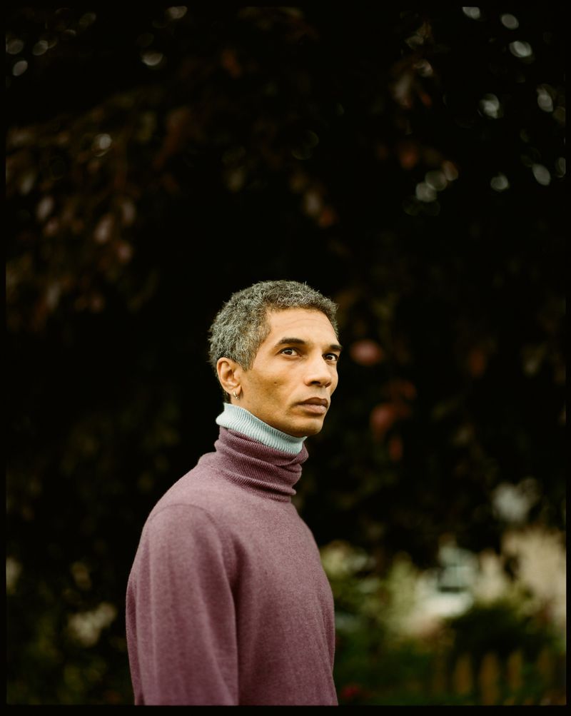 A middle-aged man with graying hair wearing a plum turtleneck sweater is pictured outdoors against a dark, leafy background.