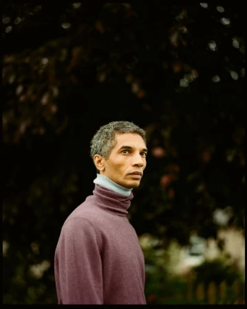 A middle-aged man with graying hair wearing a plum turtleneck sweater is pictured outdoors against a dark, leafy background.