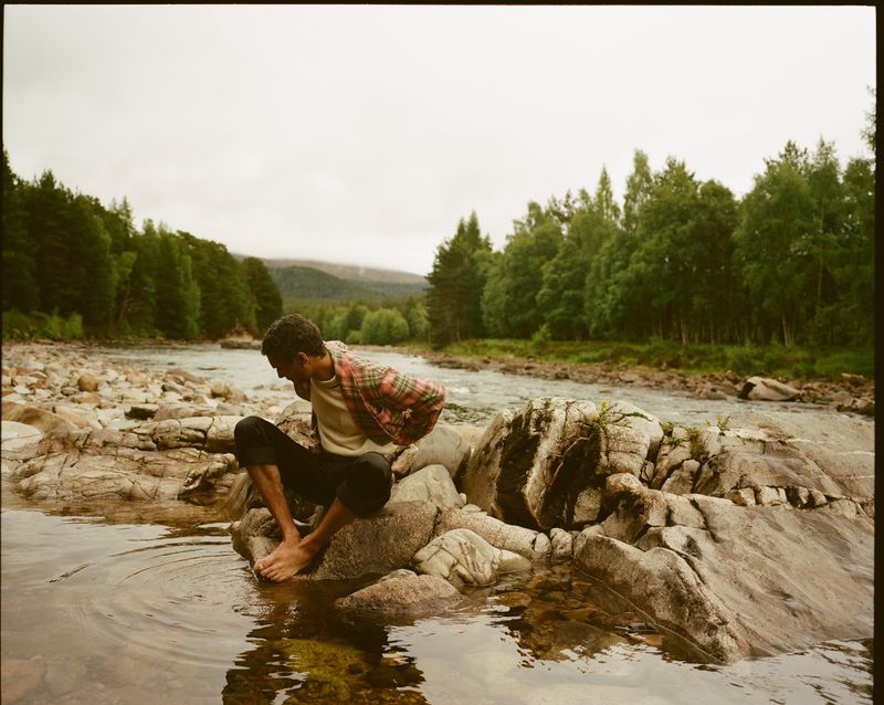 Man sits on rocks in a river wearing a red flannel shirt, captured in an outdoor setting with lush trees and a cloudy sky backdrop.