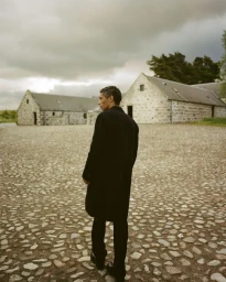 Man wearing a black coat standing on a cobblestone courtyard with stone buildings in the background under an overcast sky.