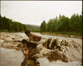 Model Filip Roseen sitting barefoot on river rocks dipping his feet into clear water surrounded by forest trees.