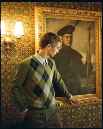 Filip Roseen wearing a green argyle sweater poses beside an ornate framed portrait against patterned wallpaper.