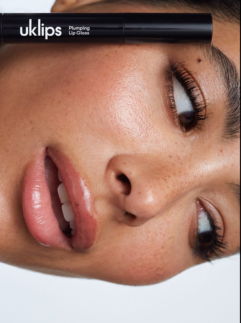 Close-up of a woman's face with clear skin and full lips. A tube of UK Lips Plumping Lip Gloss is positioned above her right eye.