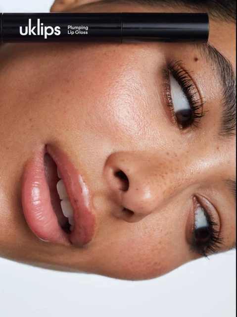 Close-up of a woman's face with clear skin and full lips. A tube of UK Lips Plumping Lip Gloss is positioned above her right eye.