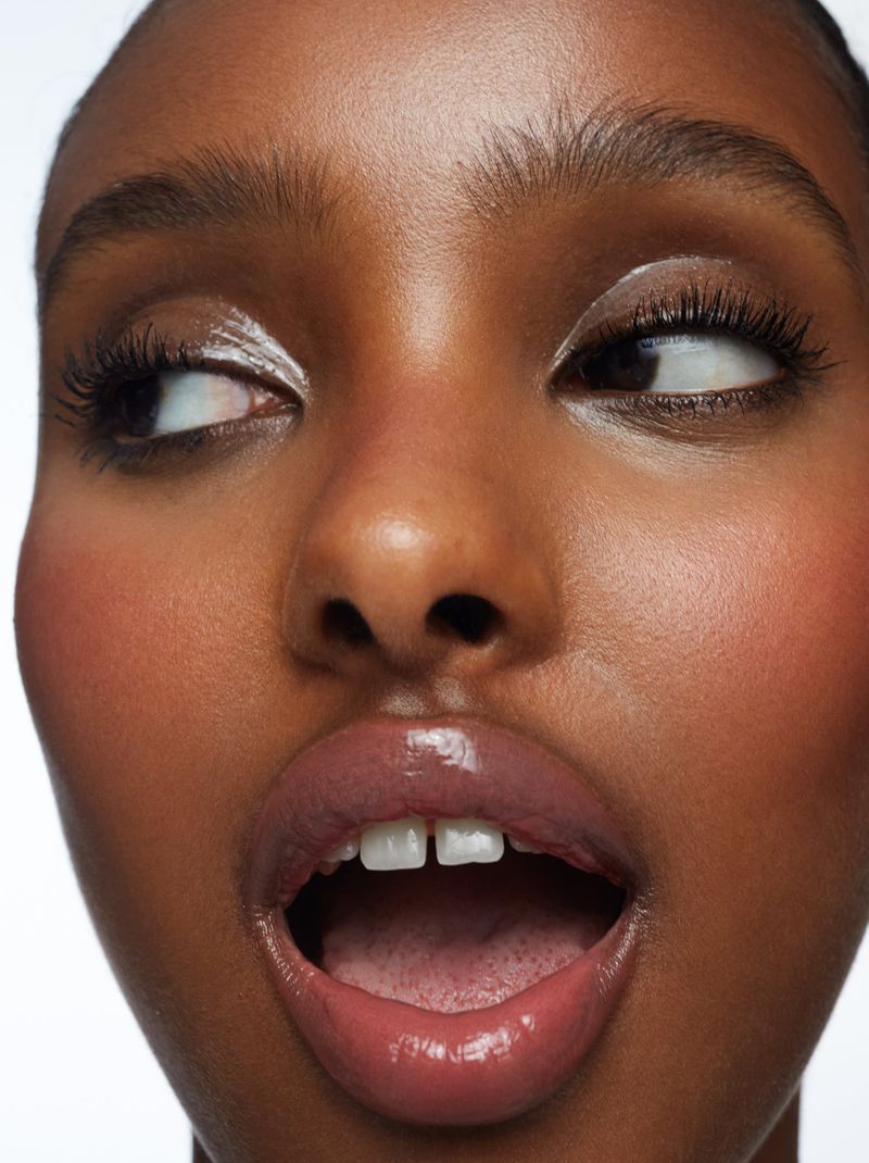 Close-up portrait of a young adult woman with an open mouth and glossy lips against a clean, white background in a beauty context.