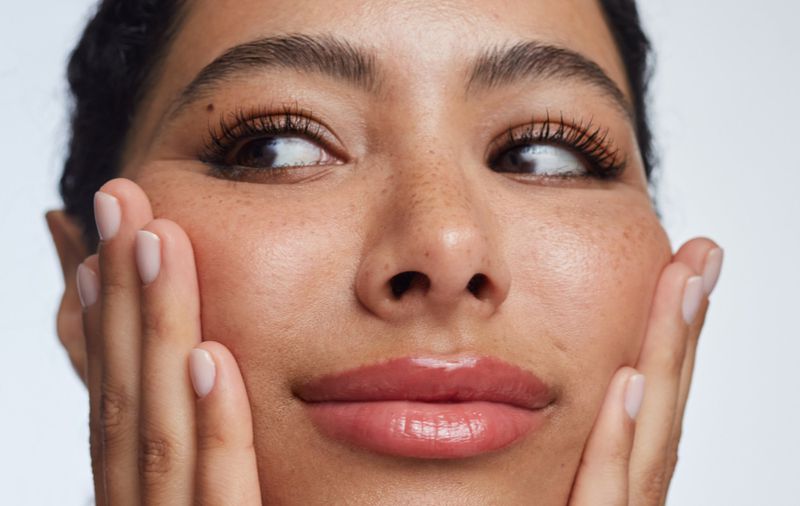 Close-up of a woman's face with her hands touching her cheeks, showing her clear skin, freckles, long eyelashes, and glossy lips.