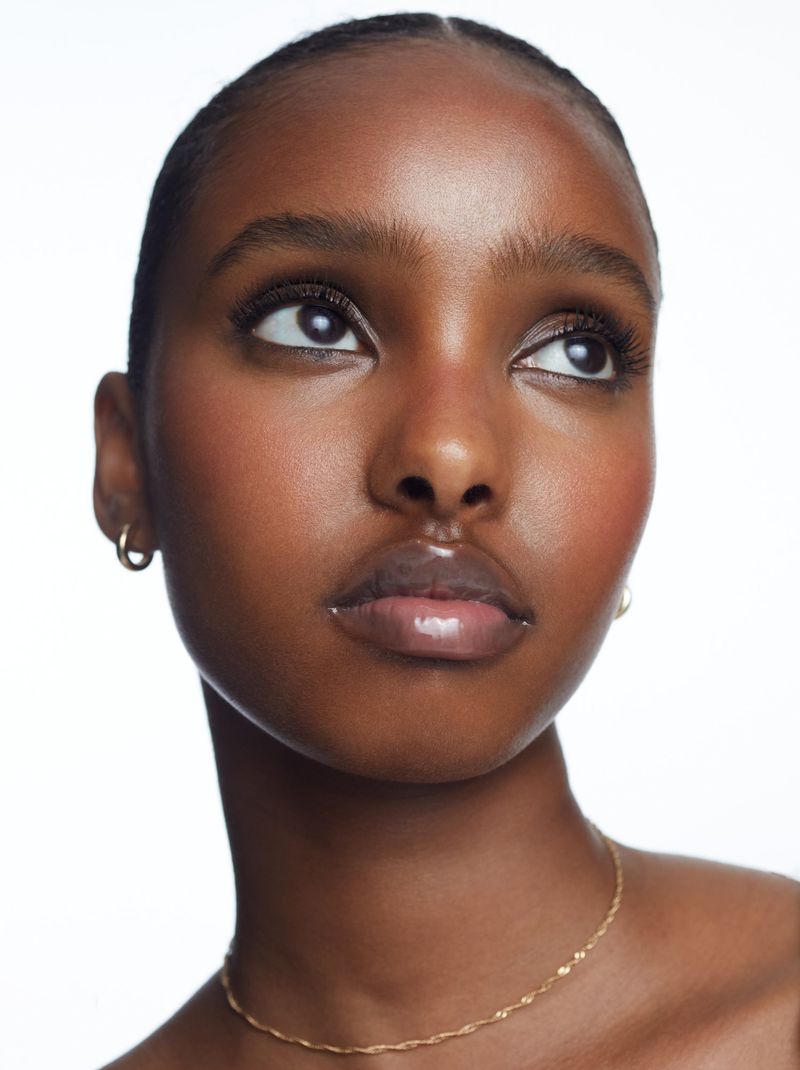Close-up portrait of an adult woman with dark skin and an elegant gold necklace, set against a clean white backdrop in a beauty style.