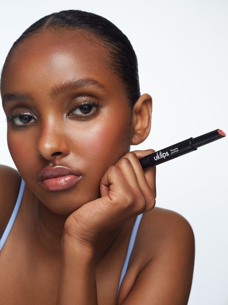 Headshot of a dark-skinned young woman holding a lipstick, set against a white background, with soft lighting emphasizing her facial features.