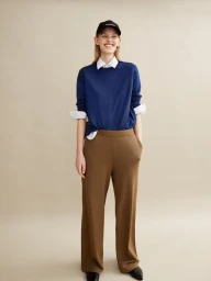 Smiling female model in a blue sweater, white collared shirt, and brown trousers standing against a seamless background
