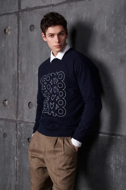 Portrait of a young adult man with dark curly hair wearing a navy sweater and khaki pants, standing against a textured grey wall indoors.