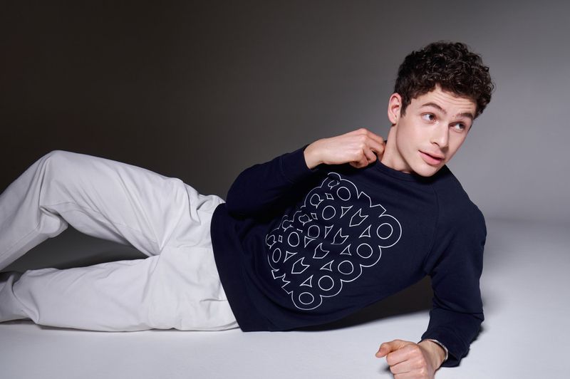 Young adult male model lies on a grey surface wearing white pants and a navy sweatshirt with a white geometric pattern. 