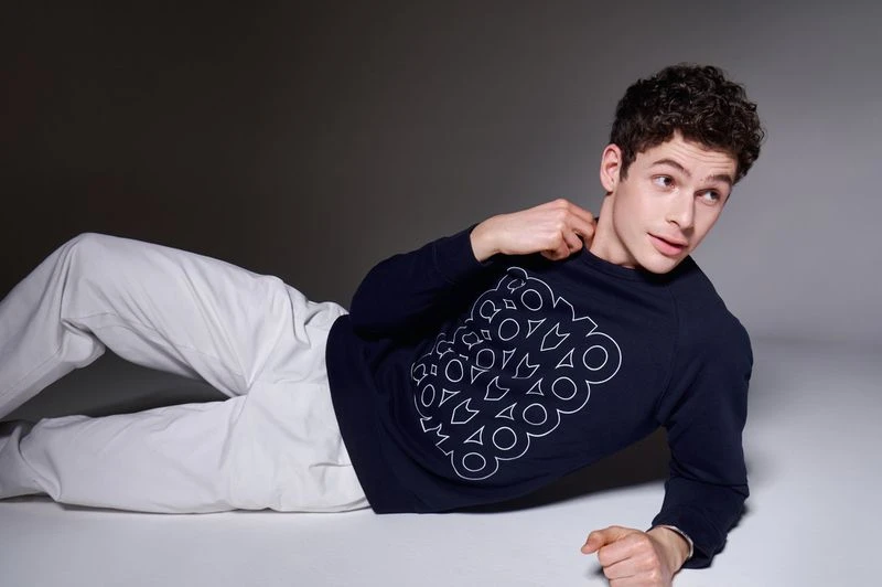 Young adult male model lies on a grey surface wearing white pants and a navy sweatshirt with a white geometric pattern. 
