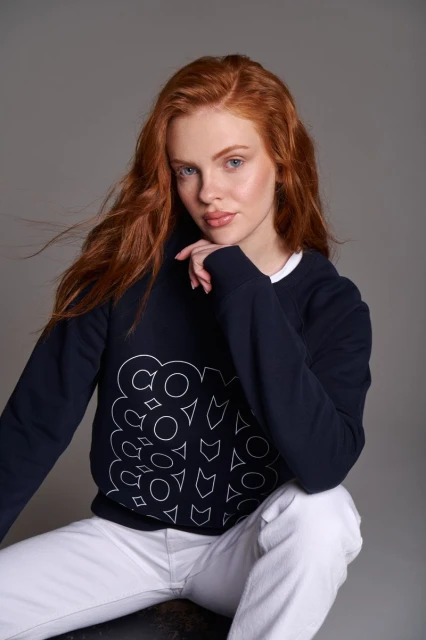 Attractive red-haired woman in a dark blue sweatshirt and white jeans sits in a studio, facing the camera, holding her hand to her face.