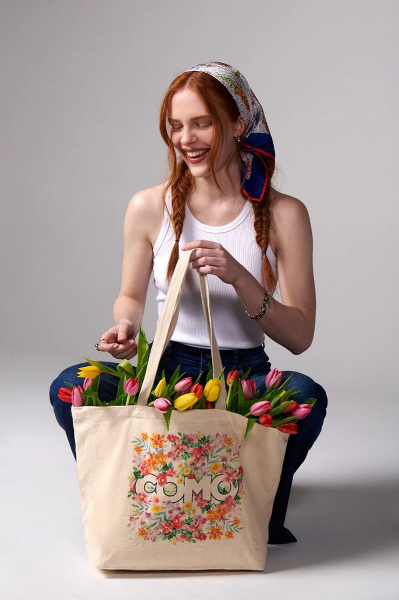 Red-haired woman smiles while holding a tote bag filled with colorful tulips. She wears a headscarf and jeans in a bright indoor setting.