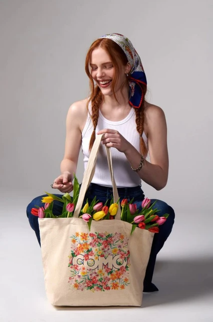 Red-haired woman smiles while holding a tote bag filled with colorful tulips. She wears a headscarf and jeans in a bright indoor setting.