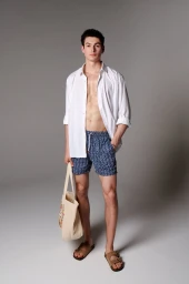 Male model in patterned blue swim shorts and unbuttoned white shirt holding a canvas tote bag against a grey seamless background