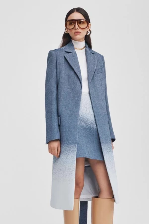 A woman wears a blue tweed coat with a white ombre effect and accessorizes with oversized sunglasses and hoop earrings in a studio shot.