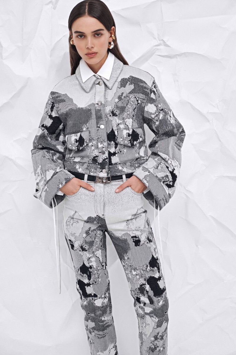 Full shot of a female fashion model wearing a grey-toned patterned denim jacket with a white collared shirt and matching jeans, set against a white backdrop.