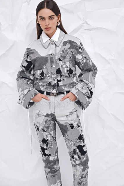 Full shot of a female fashion model wearing a grey-toned patterned denim jacket with a white collared shirt and matching jeans, set against a white backdrop.