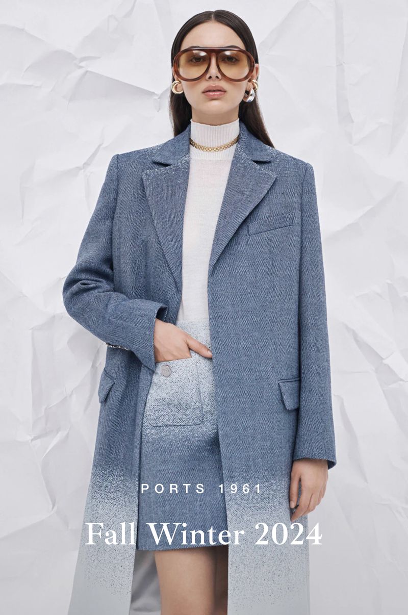 Fashion model wears a blue coat, skirt, gold jewelry, and oversized sunglasses, posing against a textured white paper background.