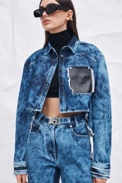 A woman in a blue acid-wash denim jacket and jeans, wearing sunglasses, poses against a white background in a fashion shot.