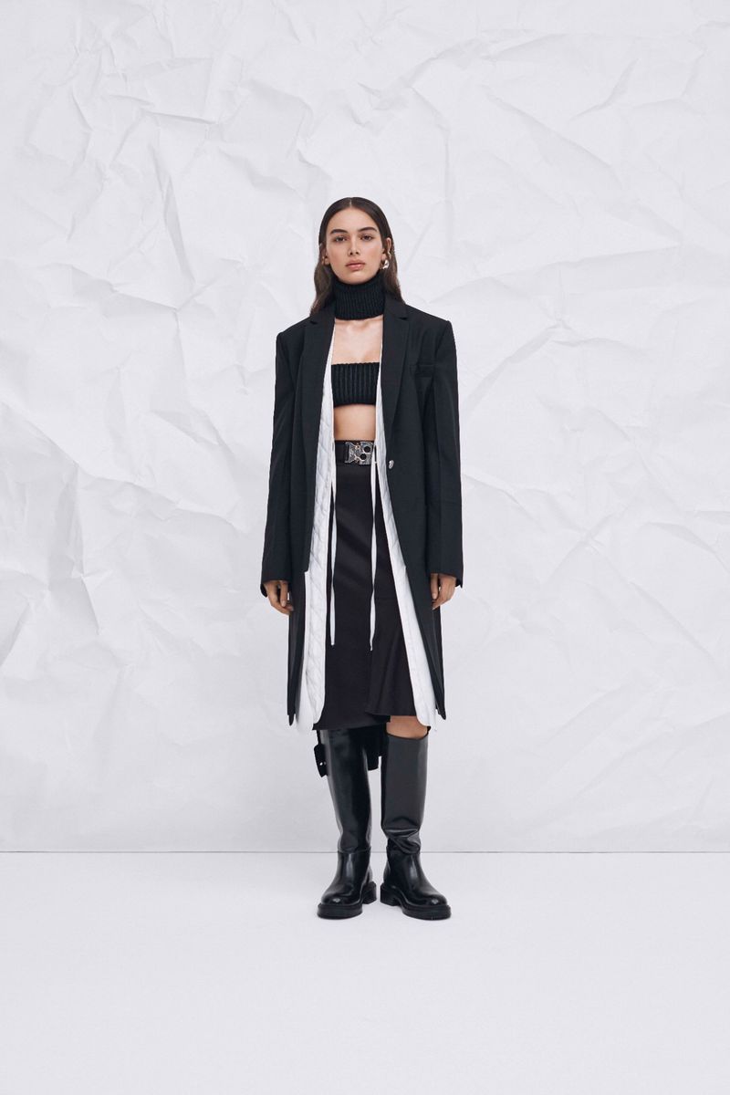 Fashion shot of an adult female wearing a black blazer, skirt and knee-high boots, posing against a white creased background.